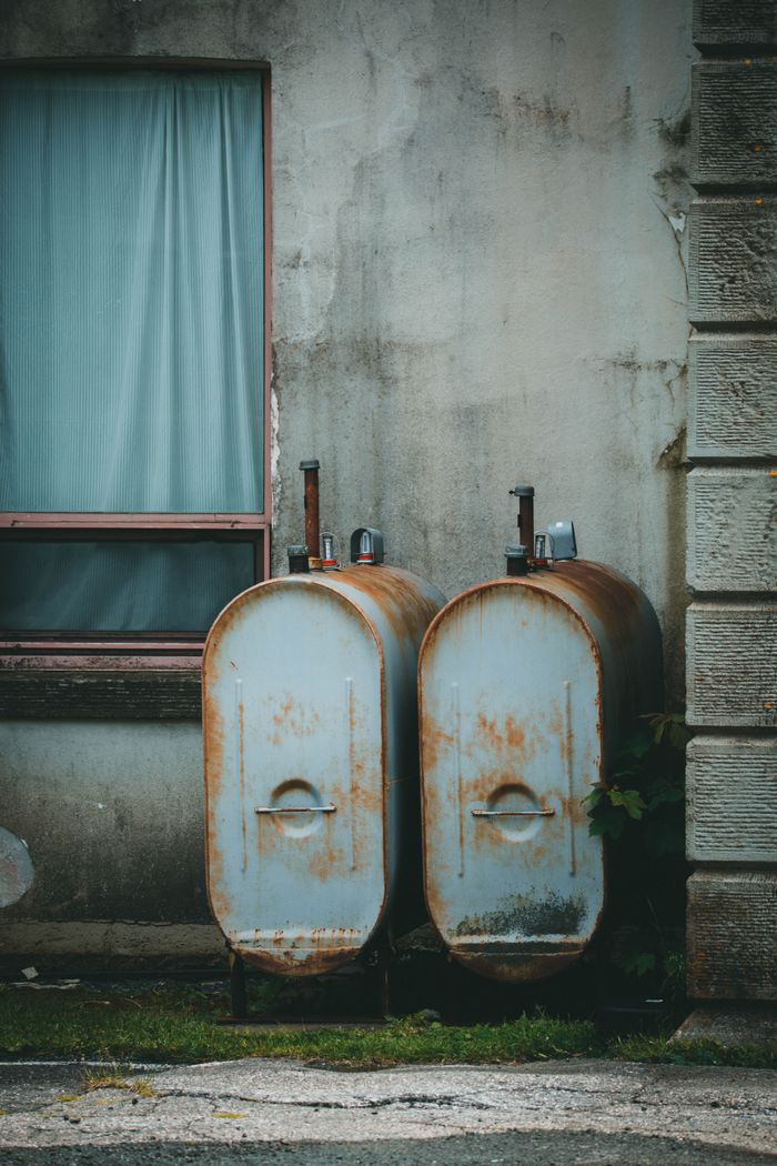 A pair of rusty tanks and a weathered building wall with a closed window, showcasing urban decay.